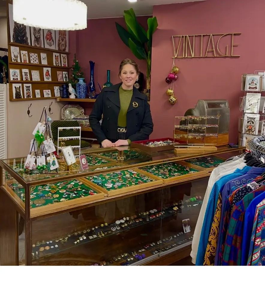 De eigenaresse van VintageGlitch achter de antieke glazen toonbank met vintage broches en sieraden