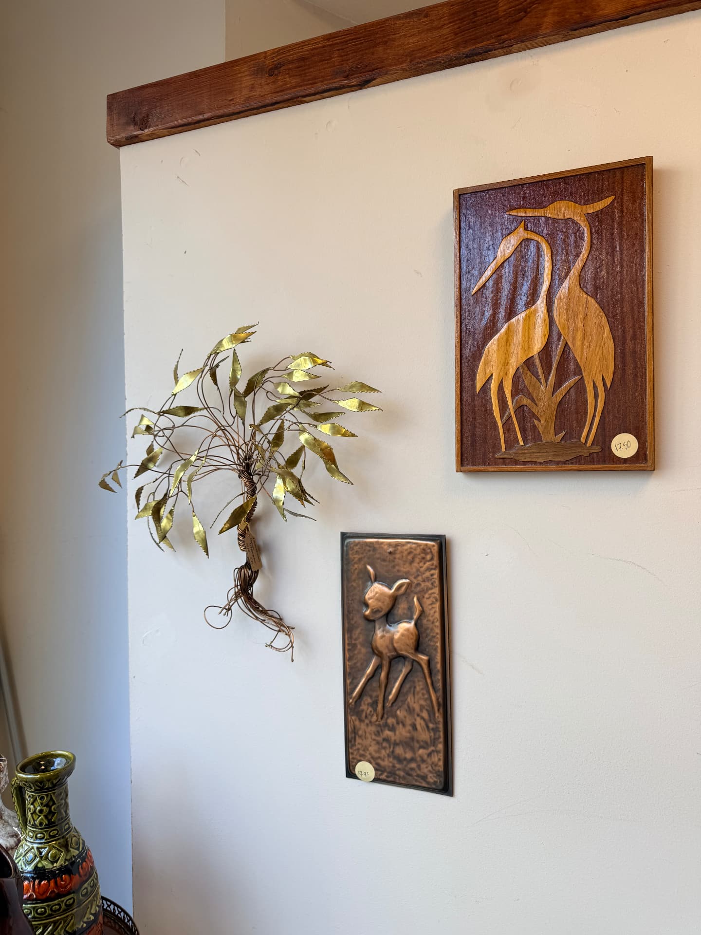 Vintage wanddecoratie aan de witte muur: een messing boompje, een houten relief met reigers en een koperen reliëf van een hertje