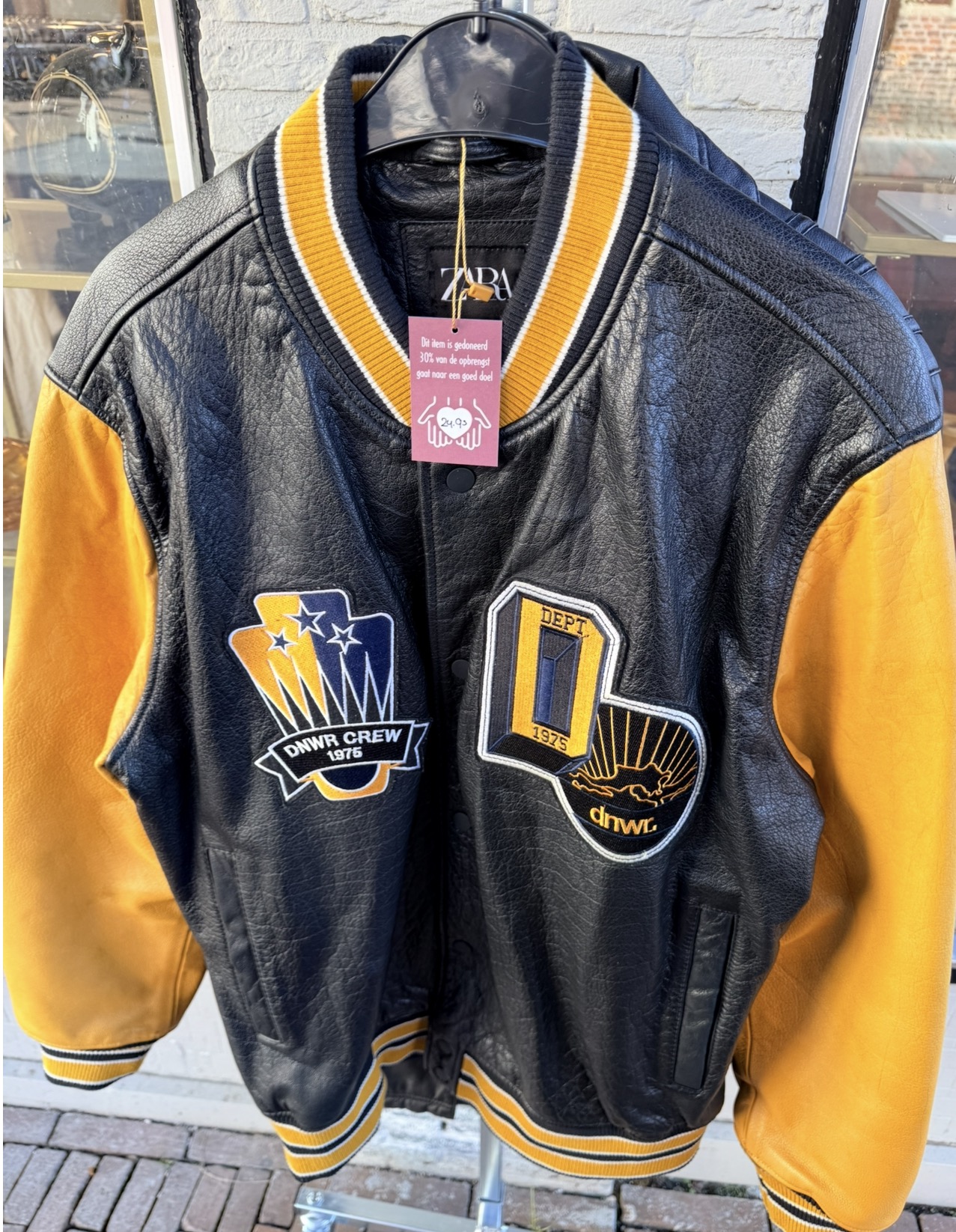 Zwart-gele leren collegejas van Zara aan een hanger buiten de winkel, met het bordeauxrode kaartje “Dit item is gedoneerd, 30% van de opbrengst gaat naar een goed doel”