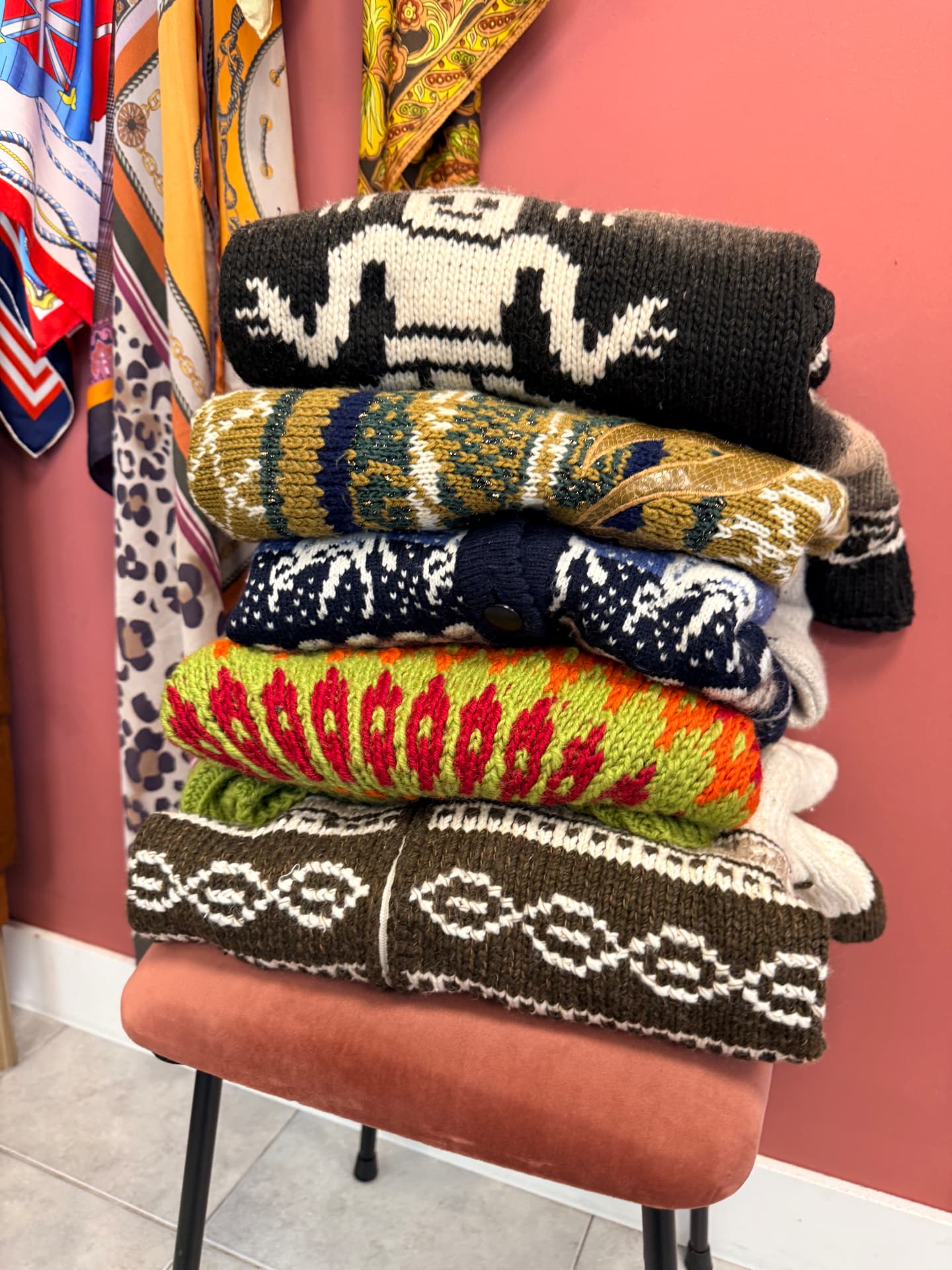 Stapel vintage gebreide truien met Noorse patronen in groen, bruin, blauw en oker op een roze velvet krukje