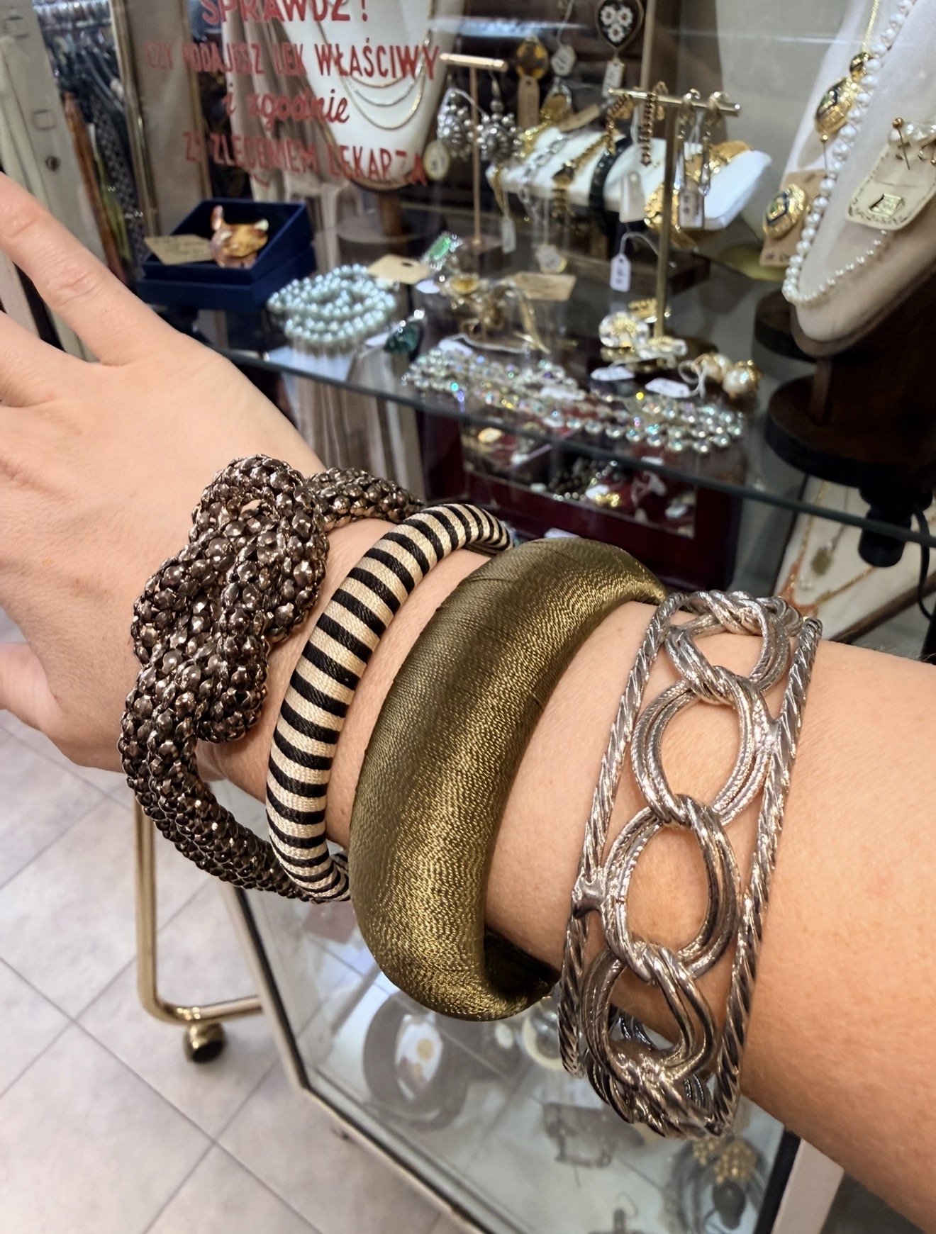 Arm met gestapelde vintage armbanden in zilver, goud en zwart-wit voor de glazen sieradenvitrine