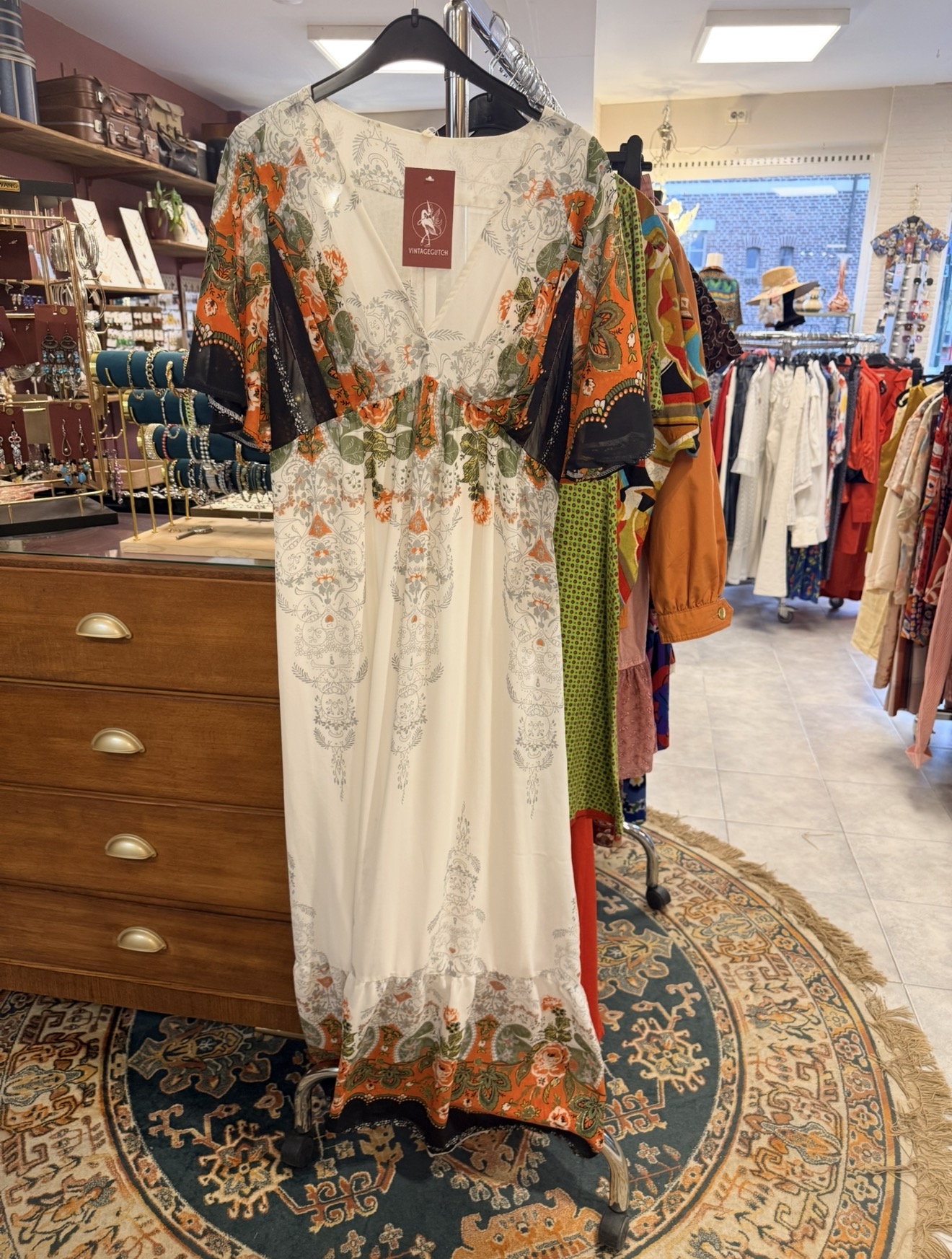 Lange crème vintage maxi-jurk met oranje en groene bloemenprint aan een rek in de winkel
